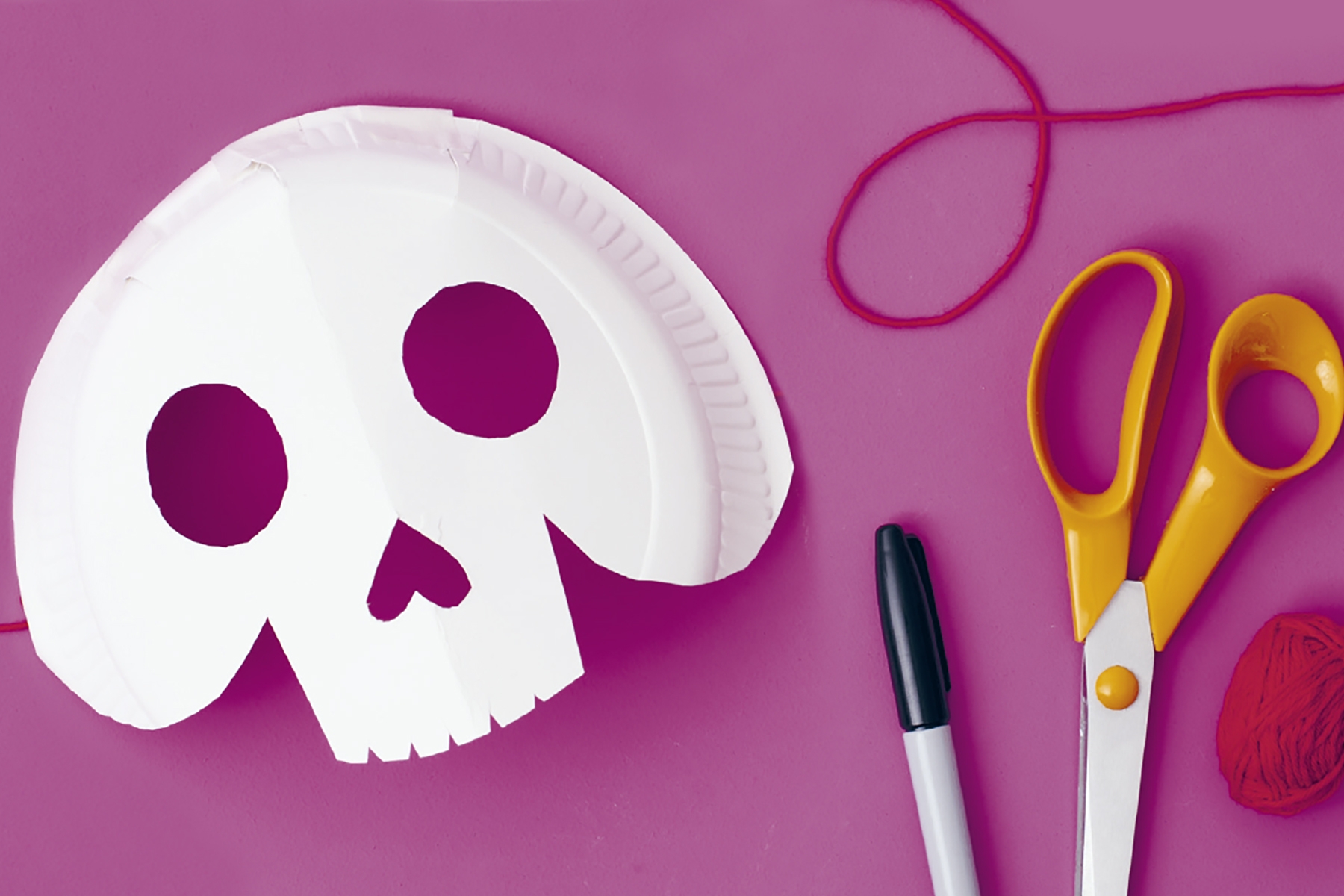A photo of a paper plate DIY skeleton mask next to some scissors, a pen and string on a magenta coloured background