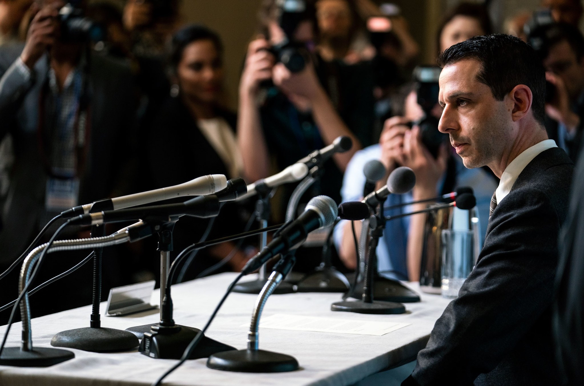 A still from the season two finale of Succession, showing Jeremy Strong as Kendall Roy seated, surrounded by bright lights and microphones at a press conference.