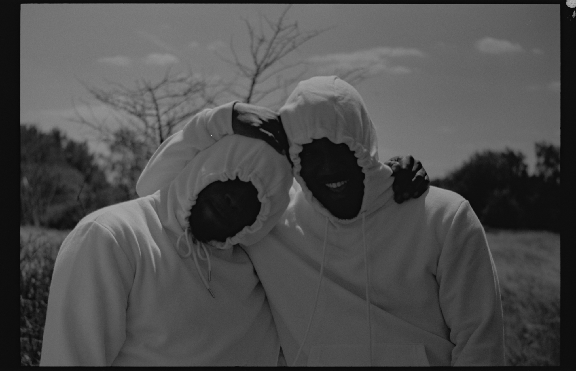 'joy, a backbone' by Caleb Femi. A black and white photograph of two men, with the hoods of their hoodies pulled up, standing with their arms around each other's shoulders.
