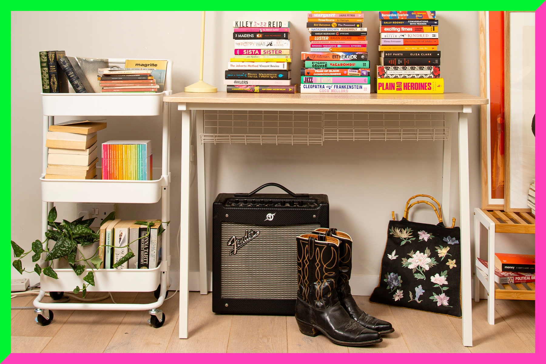 A photograph of a books trolley with books on it next to a table stacked with books. Beneath the table is a Fender amp, a pair of cowboy boots and an embroidered bag.