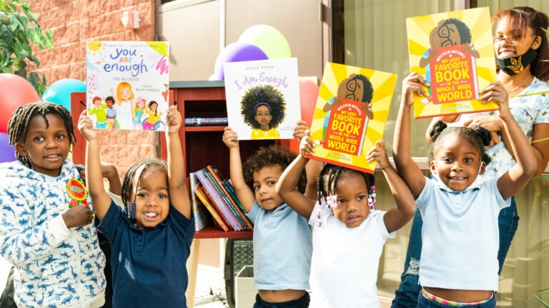 A photo of 5 children and an adult proudly holding up books in front of a Little Free Library.